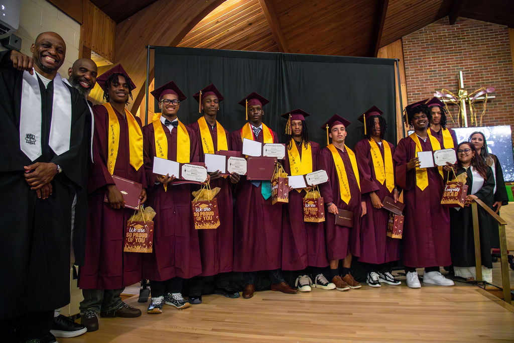 OSL graduates in maroon gowns and gold stoles holding their diplomas on stage with faculty
